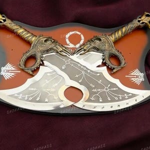 May include: A pair of silver-colored blades with intricate designs, mounted on a red and black wooden plaque. The blades have gold-colored handles with dragon-like heads.