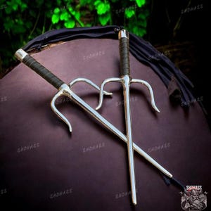 May include: Two silver sai weapons with black wrapped handles are crossed against a dark background. The sai have a three-pronged design, with a central spike and two curved side prongs. The handles are wrapped in a textured black material.