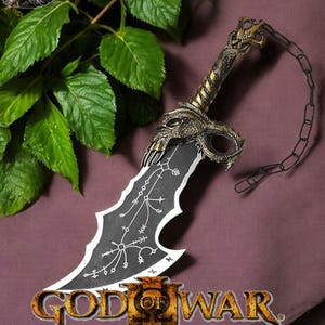 May include: A black and gold God of War III sword replica with a chain attached to the handle. The sword has a skull-like design on the handle and a black blade with white markings. The sword is lying on a purple surface with green leaves in the background.