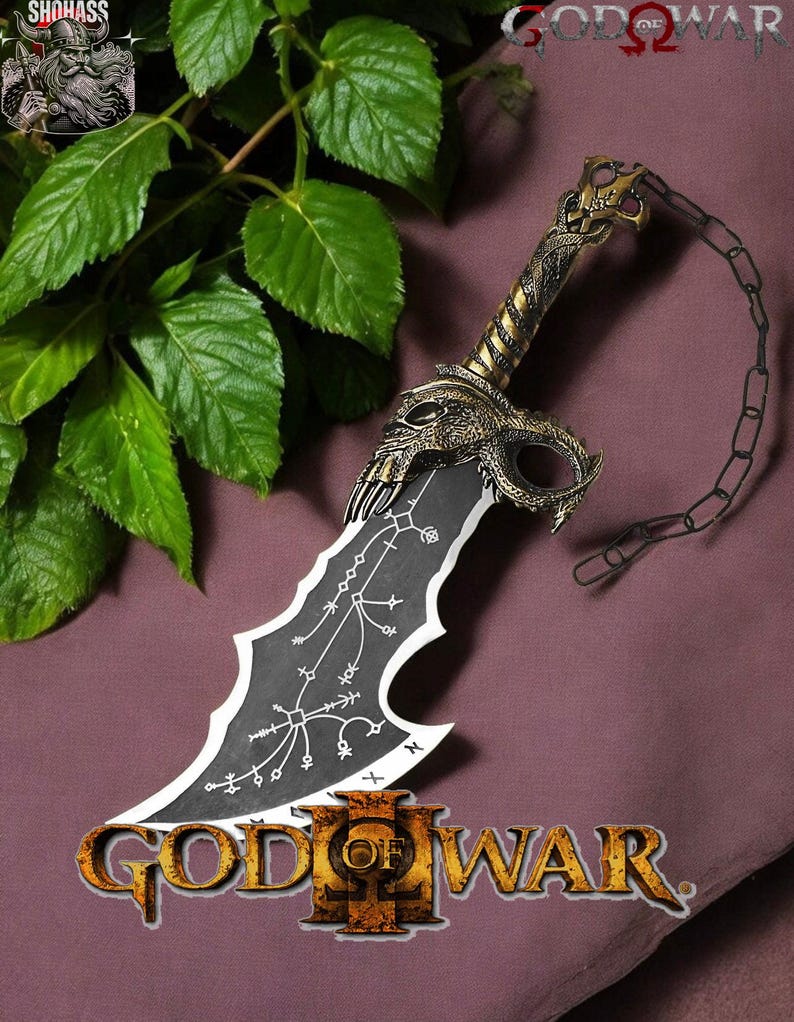 May include: A black and gold God of War III sword replica with a chain attached to the handle. The sword has a skull-like design on the handle and a black blade with white markings. The sword is lying on a purple surface with green leaves in the background.