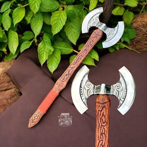 May include: A double-bladed axe with a wooden handle and a polished silver blade. The handle is intricately carved with a reddish-brown finish and wrapped with orange leather near the base. The axe is displayed against a backdrop of green leaves and brown fabric.