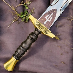 Pode incluir: Uma espada decorativa com uma lâmina prateada e um cabo castanho e dourado. A lâmina apresenta um símbolo de tridente e a palavra "ANAKAYEMOE". A espada está sobre um fundo roxo.