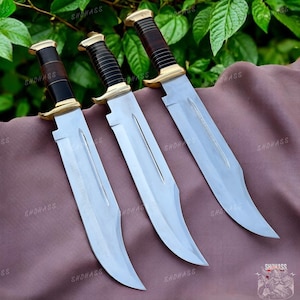 May include: Three fixed-blade knives with silver-toned steel blades, brass-coloured guards, and dark brown, ribbed handles. Each knife features a curved blade design with a central fuller groove, arranged side-by-side on a draped fabric surface.