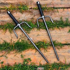 May include: A pair of black metal sai weapons featuring long, pointed central prongs and two curved side guards. Each handle is wrapped in black material, and the metal surfaces have a polished finish. The text SHOHASS is visible at the bottom.