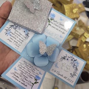 May include: A blue and silver explosion box with a butterfly design. The box is decorated with flowers and the text "15 de Agosto del 2023".