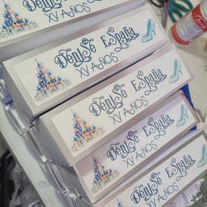May include: A stack of gift boxes with a light blue and white design. Each box features the text "Denise España XV Años", a castle illustration, and a high-heeled shoe graphic.