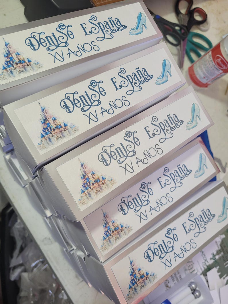 Op de afbeelding: Een stapel cadeauverpakkingen met een lichtblauw en wit ontwerp. Elke doos heeft de tekst "Denise Espa&ntilde;a XV A&ntilde;os", een kasteelillustratie en een afbeelding van een pumps.