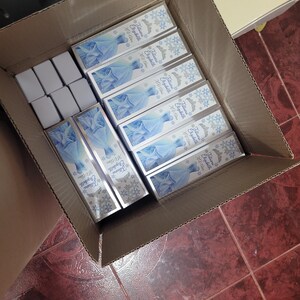 Op de afbeelding: Een kartonnen doos gevuld met rechthoekige dozen. De dozen hebben een lichtblauw ontwerp met een jurk en de tekst "Jaliana Elizabeth XU Class". Er zijn ook verschillende kleinere witte dozen zichtbaar.