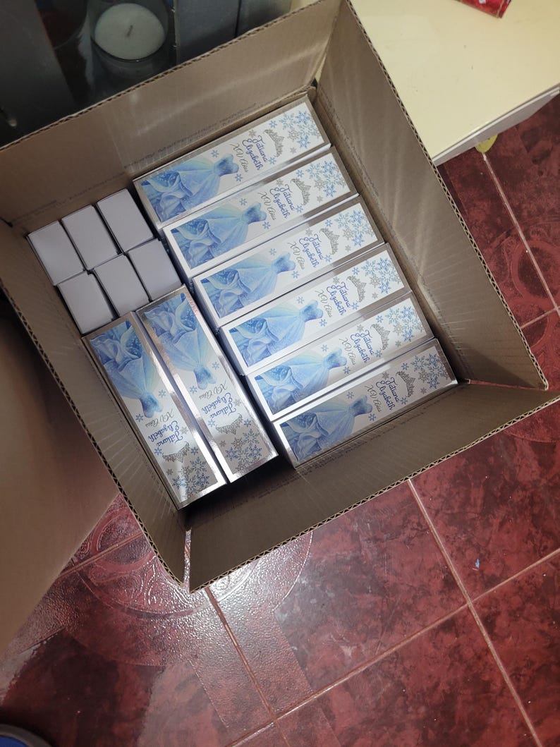 Op de afbeelding: Een kartonnen doos gevuld met rechthoekige dozen. De dozen hebben een lichtblauw ontwerp met een jurk en de tekst "Jaliana Elizabeth XU Class". Er zijn ook verschillende kleinere witte dozen zichtbaar.