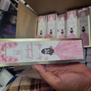 Op de afbeelding: Een rechthoekige doos met een gouden rand, met een roze bloemenontwerp met rozen en vlinders. De tekst "XV Joselyne Escobedo" wordt weergegeven. De doos wordt in een hand gehouden, met andere soortgelijke dozen op de achtergrond.