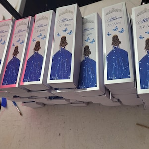 Op de afbeelding: Verschillende witte dozen met een ontwerp met een vrouw in een blauwe jurk, vlinders en de tekst "Allison XV A&ntilde;os". De dozen zijn op elkaar gestapeld.