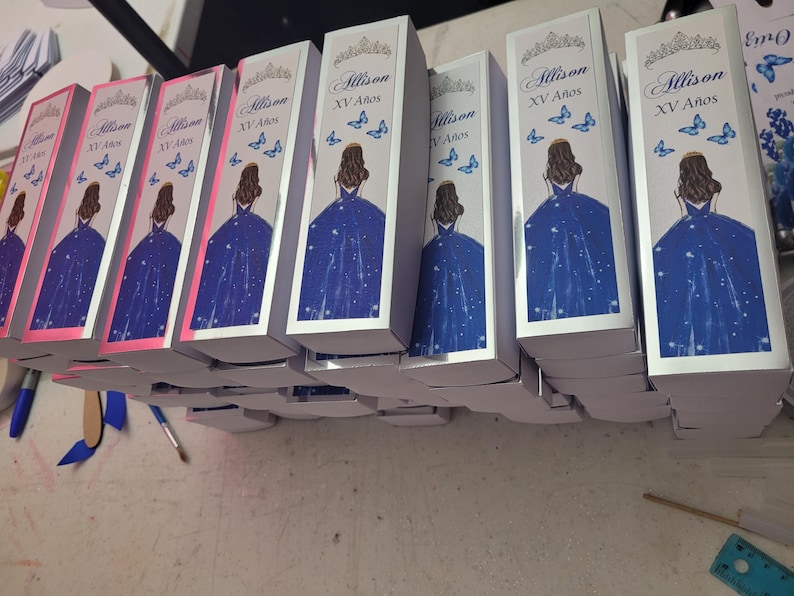 Op de afbeelding: Verschillende witte dozen met een ontwerp met een vrouw in een blauwe jurk, vlinders en de tekst "Allison XV A&ntilde;os". De dozen zijn op elkaar gestapeld.