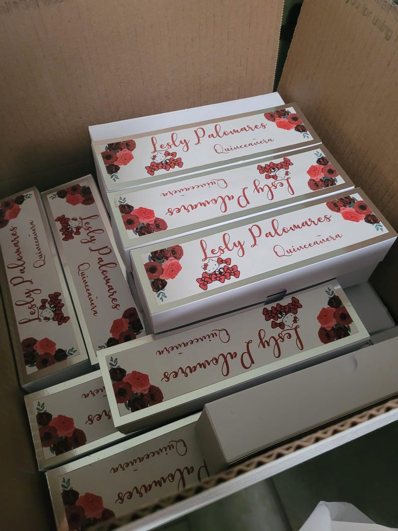 Op de afbeelding: Een kartonnen doos gevuld met rechthoekige geschenkdozen. Elke doos is wit met een zilveren rand en heeft de tekst "Lesly Palomares Quincea&ntilde;era" in rood schrift, samen met bloemen- en cartoonafbeeldingen.