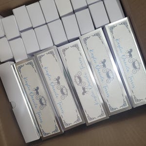 Op de afbeelding: Een doos gevuld met witte rechthoekige dozen en verschillende lange, rechthoekige dozen. De lange dozen hebben een zilveren rand en een ontwerp met een paardenkoets en de tekst "Kaylee Daniela Torres" in blauw.