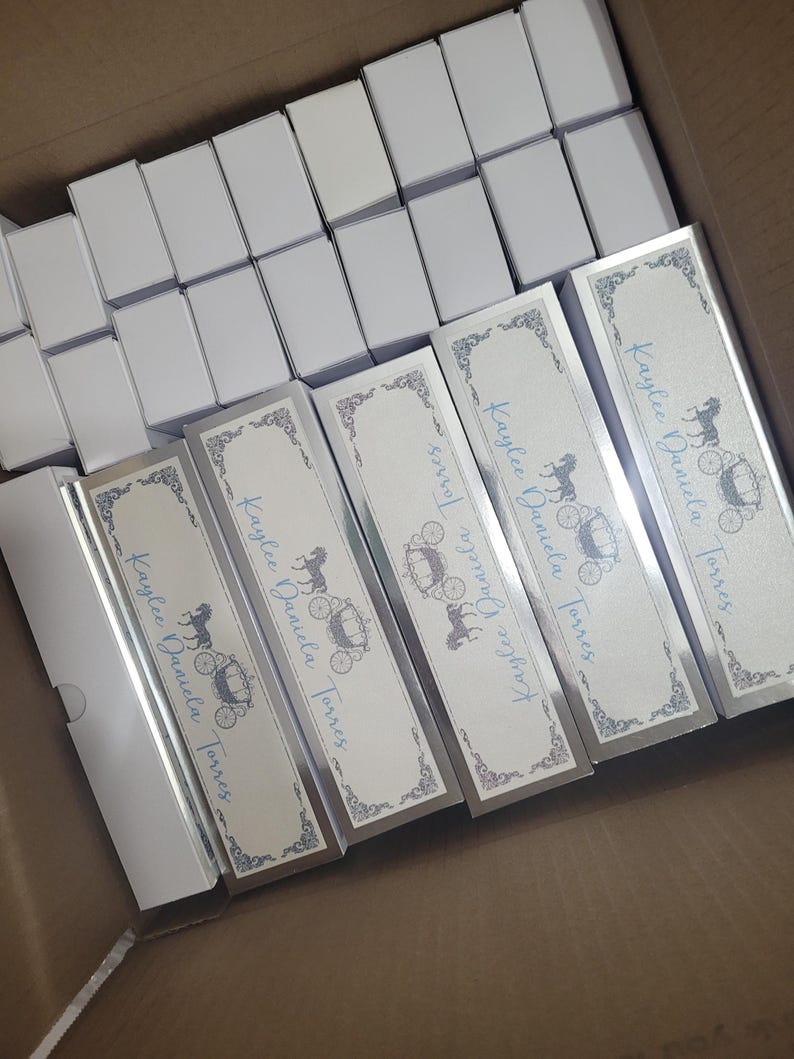 Op de afbeelding: Een doos gevuld met witte rechthoekige dozen en verschillende lange, rechthoekige dozen. De lange dozen hebben een zilveren rand en een ontwerp met een paardenkoets en de tekst "Kaylee Daniela Torres" in blauw.