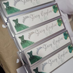 Op de afbeelding: Zilveren dozen met witte labels met een groene glitterjurk, een zilveren tiara en bloemmotieven. De tekst "Stacy Naylen XV ANOS" staat op elke doos. De dozen zijn op elkaar gestapeld.
