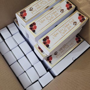 Op de afbeelding: Een kartonnen doos gevuld met witte rechthoekige dozen en verschillende rechthoekige dozen met gouden randen en bloemmotieven. De dozen met gouden afwerking hebben de tekst "Maria Jos&eacute; y Vladimir Erneste Nuestra Boda".