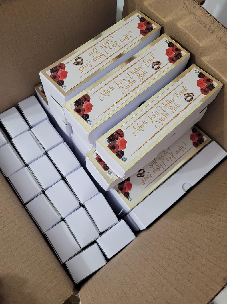 Op de afbeelding: Een kartonnen doos gevuld met witte rechthoekige dozen en verschillende rechthoekige dozen met gouden randen en bloemmotieven. De dozen met gouden afwerking hebben de tekst "Maria Jos&eacute; y Vladimir Erneste Nuestra Boda".