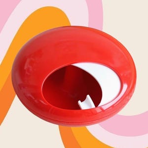 Può includere: Un contenitore rosso di forma ovale con interno bianco e apertura a forma di mezzaluna. Lo sfondo presenta un design retrò con archi rosa, arancioni e crema. L'oggetto è probabilmente in plastica.