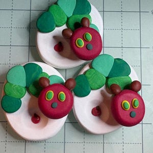 May include: Three handmade clay caterpillar figures. Each caterpillar has a red head with yellow-rimmed eyes and brown antennae. The body is made of green segments. They are on white oval bases with a red dot.