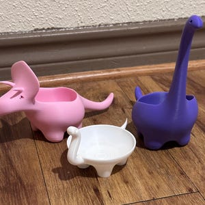 Puede incluir: Tres macetas con forma de dinosaurio en rosa, blanco y morado. La maceta de triceratops rosa tiene una abertura ancha y una cola larga. La maceta de dinosaurio blanca es pequeña y redonda. La maceta de brontosaurio morada tiene un cuello largo.
