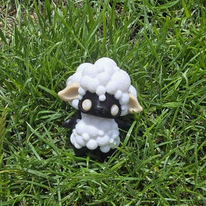 May include: A cute sheep figurine with a black face and body, white fluffy wool, and beige ears. The sheep is placed on green grass, creating a charming, pastoral scene.