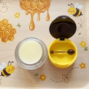 May include: A wooden tray with a honeybee theme. The tray features a jar of lip balm, a yellow container with a honey dipper, and illustrations of honeycombs, bees, and flowers. The lip balm is in a clear glass jar.