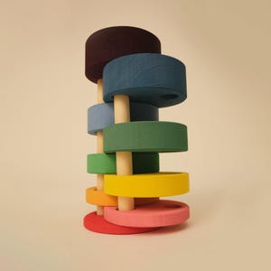 Könnte beinhalten: Ein Holz-Steckspielzeug mit sieben farbigen Ringen in Rot, Rosa, Orange, Gelb, Grün, Blau und Braun. Die Ringe sind auf einem natürlichen Holzdübel gestapelt, ein klassisches Lernspielzeug.
