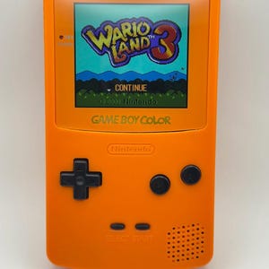 Op de afbeelding: Een oranje Game Boy Color handheld gameconsole met het spel "Wario Land 3" op het scherm. De console heeft een zwart kruis, twee zwarte knoppen en de tekst "SELECT START". Het Nintendo-logo is zichtbaar.