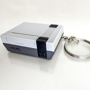 May include: A gray and white keychain shaped like a retro video game console. The keychain has a silver metal ring.