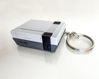 May include: A gray and white keychain shaped like a retro video game console. The keychain has a silver metal ring.