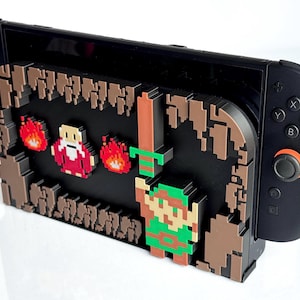 以下が含まれることがあります： 黒いNintendo Switchコンソールと、ビデオゲームシーンのピクセル化されたグラフィックを備えたカスタム3Dプリントケース。 ケースには、剣、緑色のキャラクター、炎が含まれており、すべてレトロな8ビットスタイルです。