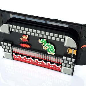 May include: Black gaming console with a pixelated Super Mario Bros. design. The design features Mario, a Koopa Troopa, and other elements from the classic video game. The console has black controllers with blue and orange accents.
