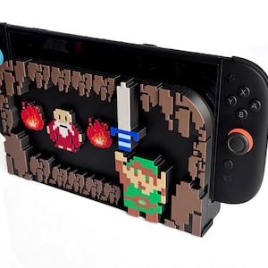 以下が含まれることがあります： 緑色のキャラクター、剣、炎、赤い姿が特徴のピクセルデザインの黒いNintendo Switchコンソール。 デザインは茶色と黒で、青とオレンジのジョイスティックが付いています。