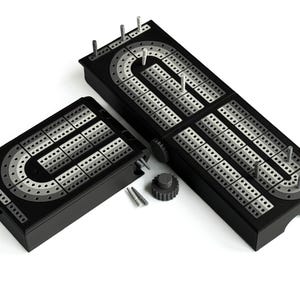 Op de afbeelding: Twee zwarte cribbageborden met zilveren en grijze scoretracks en pinnen. Eén bord is open, de andere is gesloten. Een klein, rond, zwart tandwielvormig stuk en twee zilveren pinnen zijn ook inbegrepen.