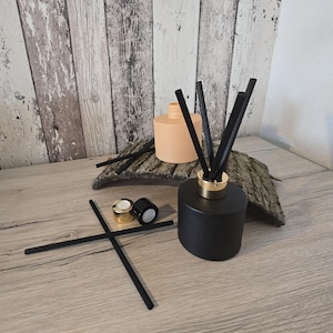 Könnte beinhalten: Ein schwarzer Diffusor für ätherische Öle mit schwarzen Stäbchen und einem goldfarbenen Kragen. Der Diffusor steht auf einer Holzoberfläche, mit einer pfirsichfarbenen Flasche und zwei Kappen im Hintergrund. Zusätzliche schwarze Stäbchen sind vorne platziert.