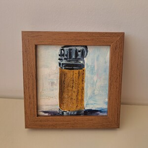 Puede incluir: Una pintura enmarcada de un encendedor amarillo con una parte superior negra. La pintura está sobre un lienzo blanco y está enmarcada en un marco de madera marrón.