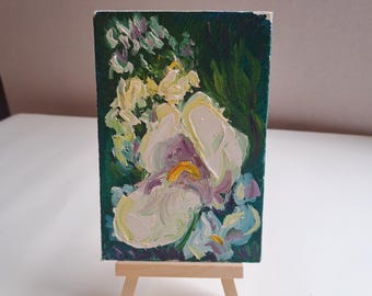 May include: A close-up of a small oil painting on canvas depicting a white flower with purple and yellow accents. The flower is surrounded by green foliage and other white flowers. The painting is on a small easel.
