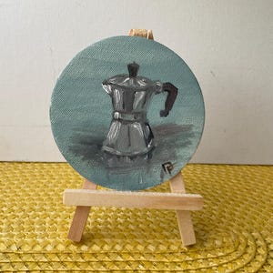 May include: A small, round painting of a silver stovetop espresso maker on a light blue background. The painting is displayed on a miniature wooden easel, set against a yellow woven surface.
