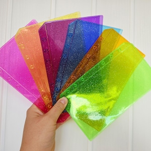 May include: A set of seven translucent, glitter-infused plastic planner dividers in a fan arrangement. The dividers feature a series of circular punched holes along one edge and come in vibrant shades of pink, orange, purple, blue, yellow, and green.