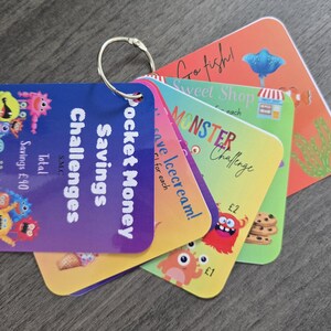 May include: A collection of pocket money savings challenge cards, held together by a metal ring. The cards feature vibrant designs with illustrations of monsters, ice cream, and other themes. Total savings amount is £40.