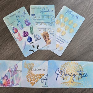 May include: A set of light blue cards with gold and blue watercolour designs. The cards feature text such as "I Am Worthy of Wealth" and "Money Tree", along with illustrations of crystals, coins, and a tree.