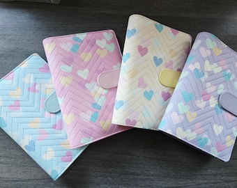 May include: Four pastel-colored notebooks with heart and chevron patterns. Each notebook has a different color: light blue, pink, cream, and lavender. The notebooks have a flap closure with a matching button.