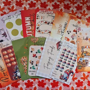 May include: A selection of autumn-themed cards with illustrations of pumpkins, leaves, and gnomes. The cards display phrases such as "Hello Autumn" and "Forging Funds." They are laid out on a surface decorated with red and orange leaf patterns.