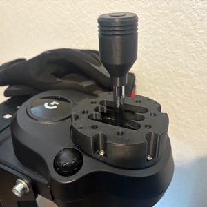 May include: A black racing simulator shifter with a cylindrical handle and a metal shaft. The shifter is mounted on a black base with a gear pattern. A pair of black gloves rests behind the shifter.