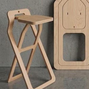 Puede incluir: Un taburete plegable de madera de color claro con un asiento cuadrado y un respaldo. El taburete tiene un diseño de patas en forma de X y está junto a un panel de madera a juego. El taburete es de madera clara.