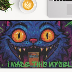 May include: A rectangular desk mat with a cartoon cat design. The cat is blue with orange eyes and a wide grin, set against a purple and green background. The text "I MADE THIS MYSELF" is in green.