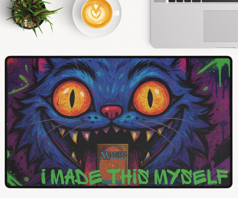 May include: A rectangular desk mat with a cartoon cat design. The cat is blue with orange eyes and a wide grin, set against a purple and green background. The text "I MADE THIS MYSELF" is in green.