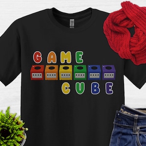 May include: Black t-shirt with the words "GAME CUBE" in colorful letters. The word "GAME" is above a row of six colorful GameCube consoles. The word "CUBE" is below the consoles. A red scarf and a small green plant are also visible.