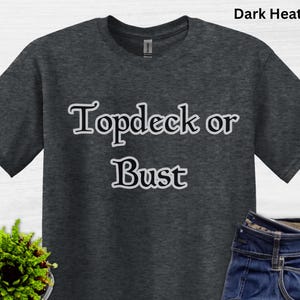 May include: Dark heather gray t-shirt with the text "Topdeck or Bust" in a white, outlined font. The shirt is paired with blue jeans. A small potted plant is visible in the lower left corner.